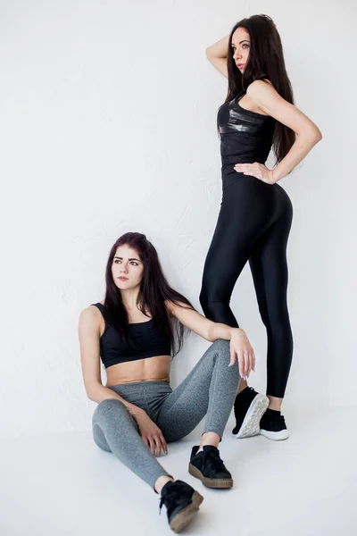 Portrait of two charming athletic sportwomen in sportsuits in white studio. One of them is leaning on the white wall and looking aside while anothe sitting on the floor and looking in camera. — Stock Photo, Image