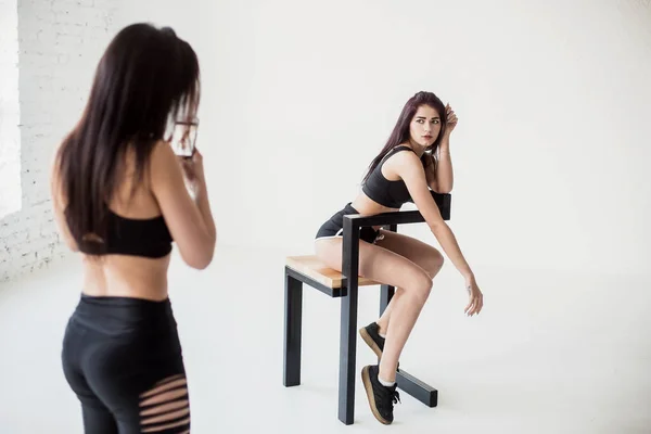 Rückansicht der sportlichen Frau beim Fotografieren ihrer athletischen Freundin in kurzen Oberteilen und kurzen Hosen, die auf dem Stuhl vor weißem Hintergrund posiert. — Stockfoto