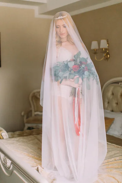 Mariée blonde avec long voile vêtue de sous-vêtements blancs sexy debout sur le lit et tenant un bouquet de mariage — Photo