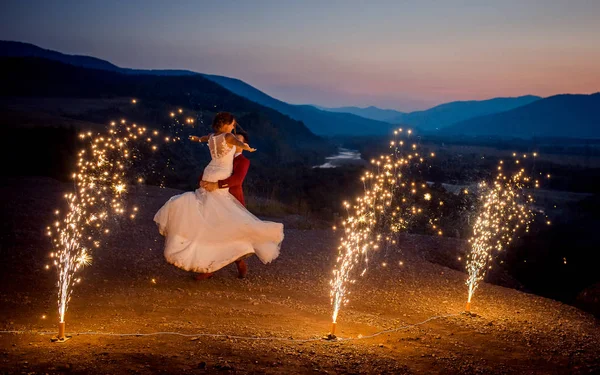 新郎がこんがらがってる花火の燃焼で飾られた山に彼の魅力的な花嫁. — ストック写真