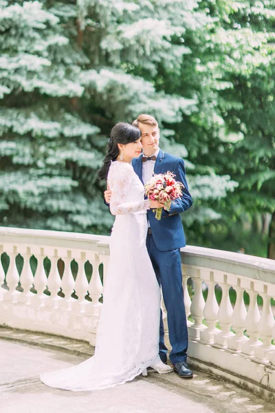 De gelukkige pasgetrouwden zijn knuffelen en opzij kijken met het boeket van de bruiloft. Volledige weergave. — Stockfoto