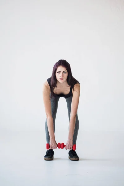 Front full-length view of the beautiful athletic woman bending with the red dumbbells in the studio. — Stock Photo, Image