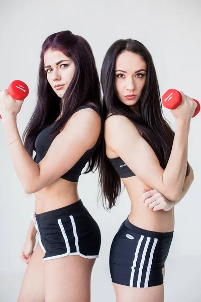 Портрет двух привлекательных спортивных женщин в шортах и коротких топах, стоящих спиной к спине с гантелями на белом фоне . — стоковое фото