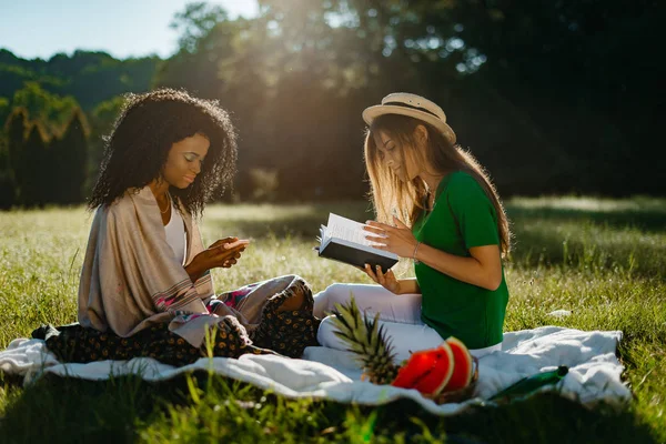 Dva multietnické dívka přátelé během pikniku na slunečné louce. Atraktivní Africká dívka je chatování a procházení prostřednictvím mobilního telefonu, zatímco její přítel hezká blondýnka je čtení knihy. — Stock fotografie