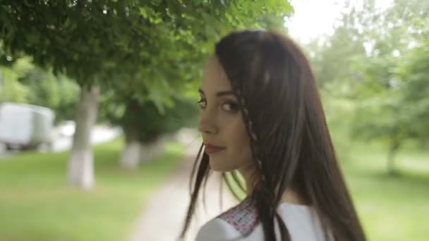 Vista posteriore dell'affascinante bruna sorridente in camicia da ricamo che si gira mentre cammina lungo il parco soleggiato . — Video Stock