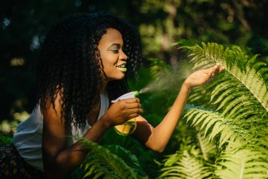 Güzel genç mutlu Afrika kız yeşil ruj ve göz gölgeler ferns için yeşil plastik tetikleyici sprey kullanırken dikkat çekici.