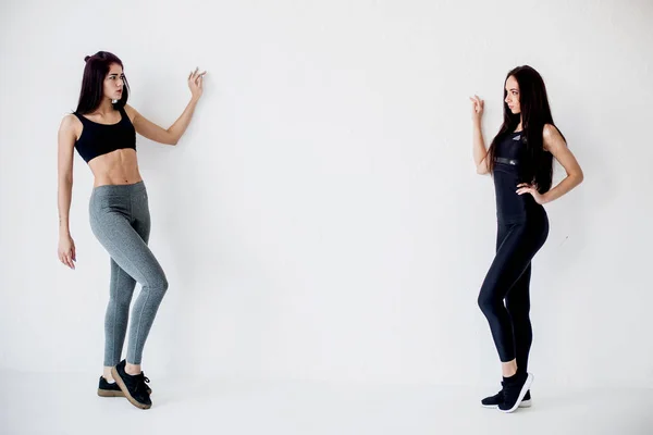 Full-length horizontal shot. Two athletic charming young sportwomen looking at each other while posing at the white wall in sportsuits.
