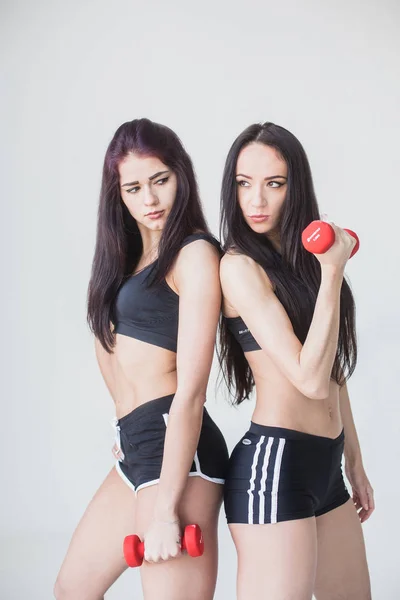 Zwei junge Sportlerinnen in Schlaghosen und kurzen Hosen posieren mit Hanteln und blicken beiseite auf weißen Hintergrund. — Stockfoto
