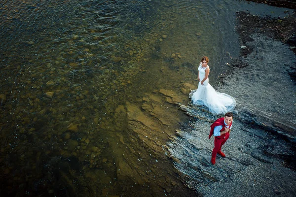 Vista acima no elegante casal recém-casado em pé na margem do rio. O noivo está a usar o fato vermelho. . — Fotografia de Stock