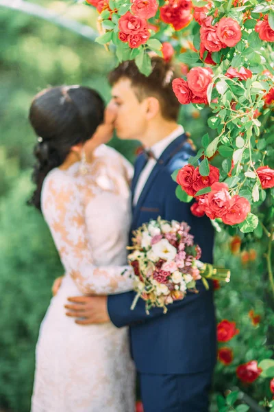 Vue rapprochée des rosiers rouges sur le fond flou des jeunes mariés qui s'embrassent . — Photo