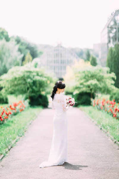 La vue arrière pleine longueur de la mariée dans la longue robe blanche tenant le bouquet de mariage dans le parc . — Photo