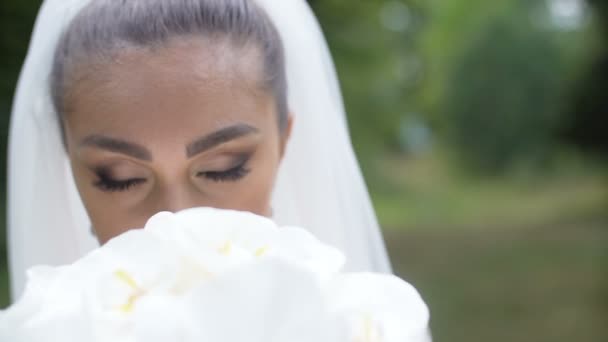 Close-up retrato da jovem noiva morena atraente cheirando o buquê de casamento de flores brancas e olhando para a câmera no fundo verde turvo . — Vídeo de Stock