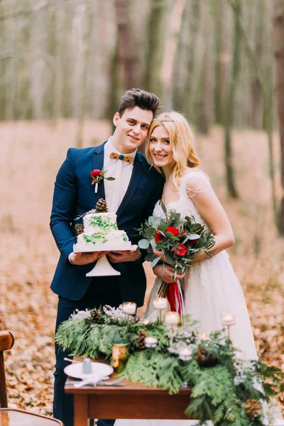 Beau couple de mariage posant avec gâteau dans les mains près de la table vintage. Fond de forêt d'automne — Photo