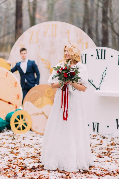 Gorgeous blonde bride with bouquet in the autumn forest. Silhouette of groom on background.