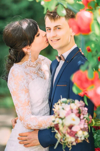 A noiva está beijando o noivo sorridente na bochecha enquanto segura o buquê de casamento . — Fotografia de Stock