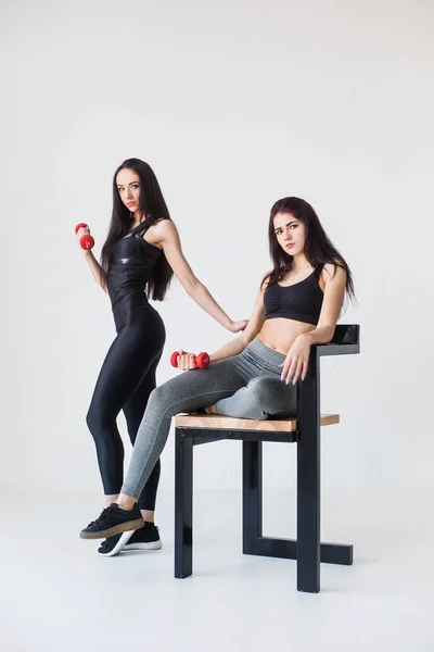 Junge muskulöse Frau sitzt auf dem Stuhl, während ihre sportliche Freundin neben ihr steht. Sie halten die Hanteln vor dem weißen Hintergrund. — Stockfoto
