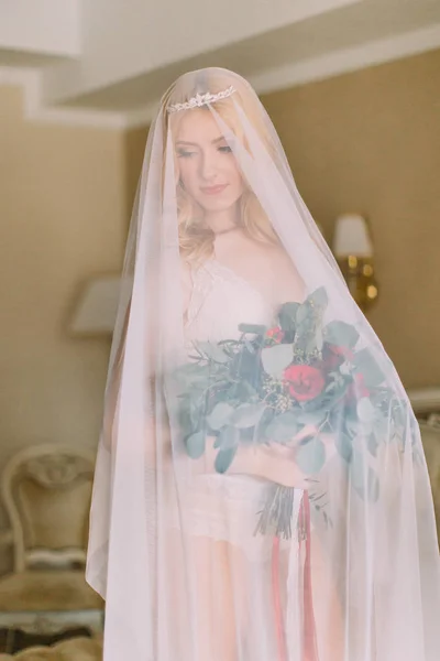 Blonde bruid met lange sluier gekleed in sexy wit ondergoed permanent op het bed en het bedrijf van de bruiloft boeket — Stockfoto