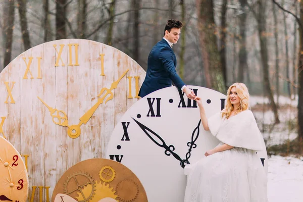 Feliz pareja de boda suavemente cogidos de la mano en los grandes relojes vintage en el bosque de otoño. Decoraciones creativas — Foto de Stock