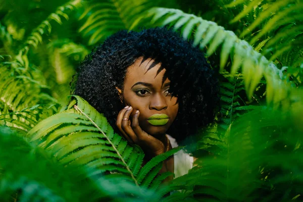 Горизонтальный портрет. Довольно привлекательная африканская девушка с зеленой помадой и тенями для век нежно прикасаясь к ее лицу в окружении папоротников . — стоковое фото