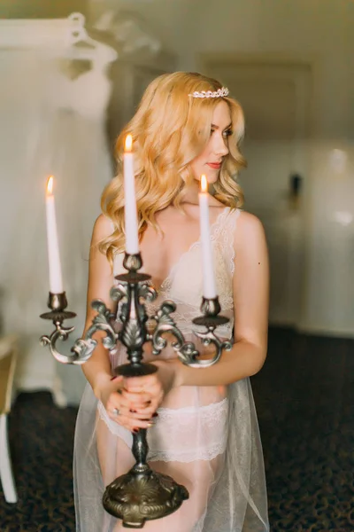 Sexy novia rubia en ropa interior blanca y diadema de diamantes en la cabeza sosteniendo viejo candelabro. Fondo de apartamentos de lujo — Foto de Stock