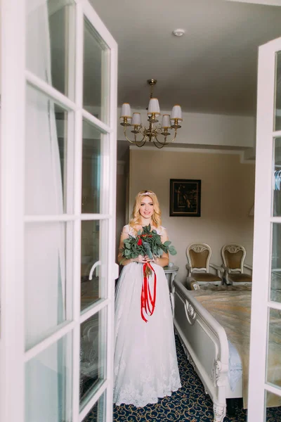 Hermosa novia rubia con ramo de rosas rojas de pie cerca de la ventana — Foto de Stock