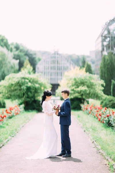La vue latérale pleine longueur du couple heureux nouvellement marié tenant le bouquet de mariage dans le parc . — Photo