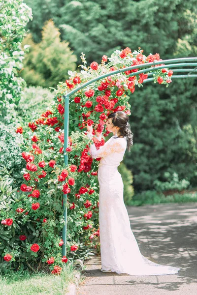 Fullängds beskåda av den bruden stående inder bågen täckt med röda rosor. Hon lukta och röra blommor. — Stockfoto