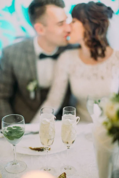 Kieliszki do szampana są umieszczane na stole ślub w niewyraźne tło Całowanie nowożeńcy. — Zdjęcie stockowe