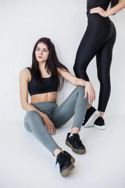 The young charming athletic brunette in sportsuits is sitting on the white floor at the background of the legs of her friend. — Stock Photo, Image