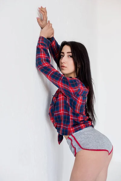 Hermosa mujer encantadora con pelo largo y oscuro en pantalones cortos y camisa de celda rosa apoyada en la pared blanca . — Foto de Stock