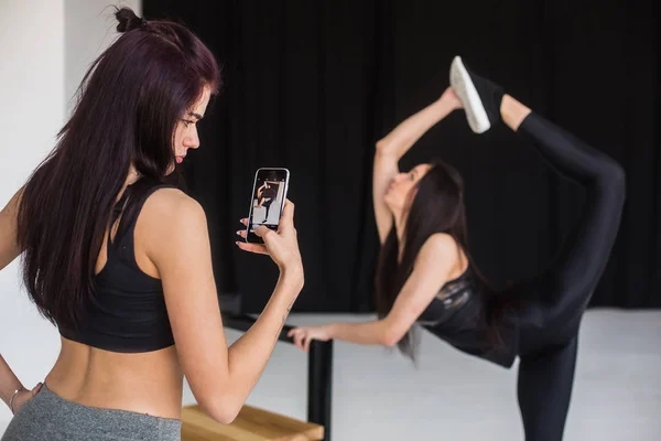 Rear view of the young athletic woman taking photo of her sportive friend in the standing bow yoga pose. View in the mobile phone. — Stock Photo, Image