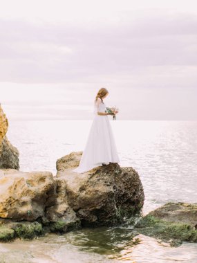 Gelin düğün buket tutarak uzun beyaz elbiseli deniz kenarında yıkanmış taş duruyor.