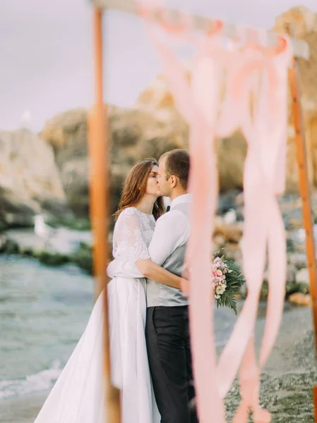 Gli sposi baciare dietro l'arco di nozze decorato con nastri rosa sullo sfondo del mare . — Foto Stock
