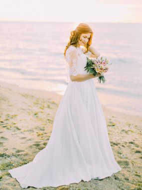 Güneşin arka planda deniz, buket tutarak uzun düğün elbise gelin yan görünüm.