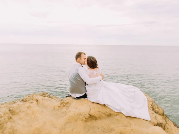 Романтический задний портрет целующейся пары, сидящей на скале на фоне моря во время заката . — стоковое фото