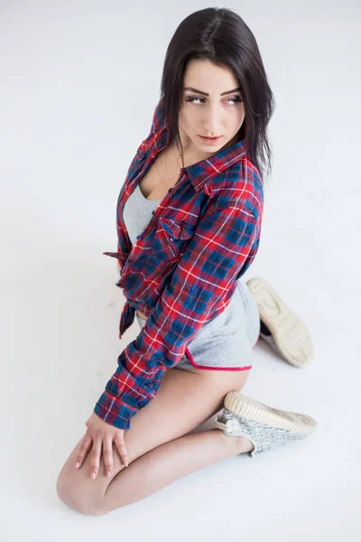 Above view of the young attractive brunette with natural make-up in short shorts and cell shirt sitting on the white floor and looking away. — Stock Photo, Image