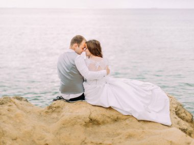 Yatay arkadan görünüm, arka plan deniz, Uçurumdaki kafa kafaya oturan güzel yeni evliler.
