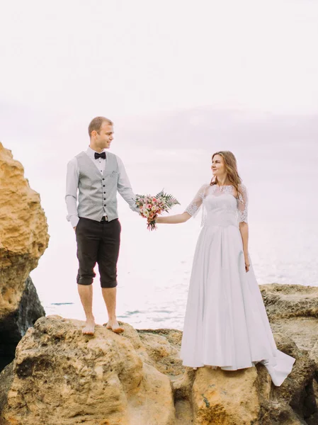 Felice sposi che si tengono per mano e si guardano mentre stanno in piedi scogliera sullo sfondo del mare . — Foto Stock