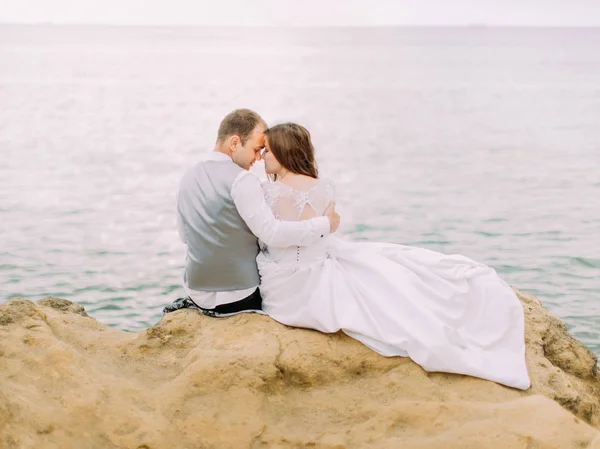 La vista trasera horizontal de los encantadores recién casados sentados cabeza a cabeza en el acantilado al fondo del mar . —  Fotos de Stock