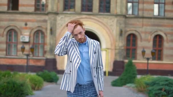 Walking stylish man with beard touching his ginger curly hair at the background of the building. He is wearing striped jacket and trews pants. — Stock Video