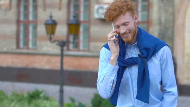 Homem sorridente feliz com cabelo encaracolado gengibre e barba conversando através do telefone celular no fundo turvo do edifício . — Vídeo de Stock