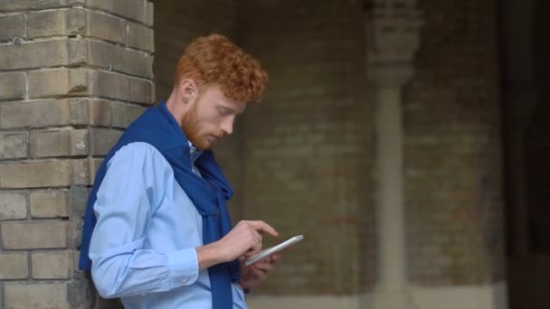 Side half-lenght portrait of the man with red ginger hair and bear chatting, browsing and texting via the mobile phone while leaning on the brick wall. — Stock Video
