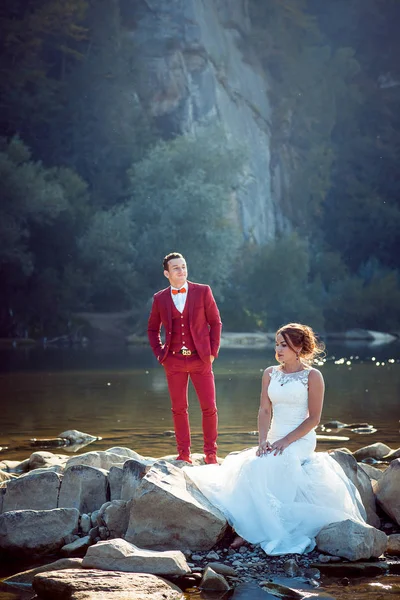 Retrato vertical de la boda de la encantadora pareja de recién casados descansando cerca del río. La novia de la cabeza roja está sentada en la piedra mientras el novio en traje rojo de pie detrás . — Foto de Stock