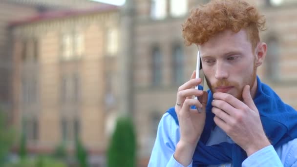 Un homme à tête de gingembre avec barbe parle via le téléphone portable sur le fond flou du bâtiment. Gros plan sur l'homme inquiet . — Video