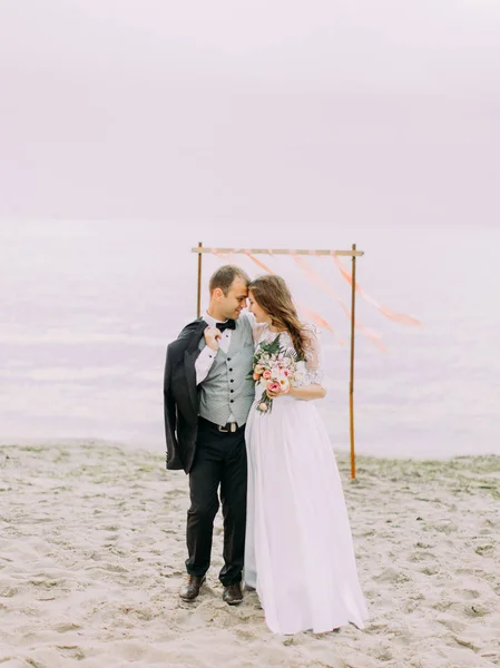 Повнометражний вигляд щасливих молодят, що стоять спереду до голови перед весільною аркою на пляжі . — стокове фото