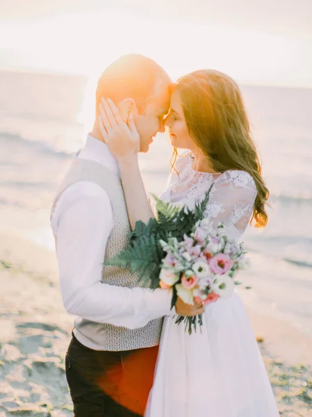 Portrait mignon du couple heureux. La mariée caresse le visage du marié sur le fond du coucher de soleil dans la mer . — Photo