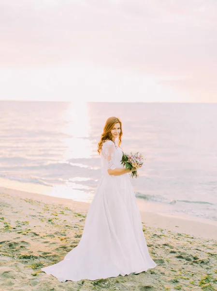 La vista completa della sposa nel lungo abito da sposa che tiene il bouquet sullo sfondo del tramonto nel mare . — Foto Stock