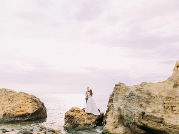 A vista de trás dos recém-casados em pé no penhasco entre o oceano durante o pôr do sol . — Fotografia de Stock