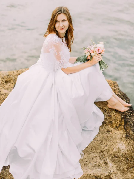 A vista de perto da noiva com o buquê sentado no penhasco e olhando para o lado direito no fundo do mar . — Fotografia de Stock