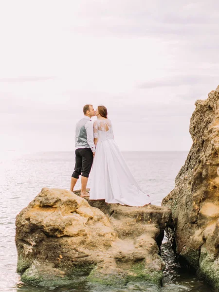 Całowanie nowożeńcy stojąc na klifie wśród morza. Widok z tyłu. — Zdjęcie stockowe